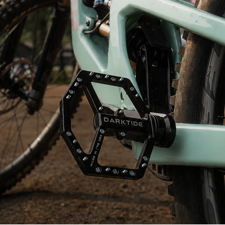 A close-up outdoor shot of a mountain bike frame and pedal area. The bike frame is a pale mint green, with a black Darktide pedal and black suspension components.